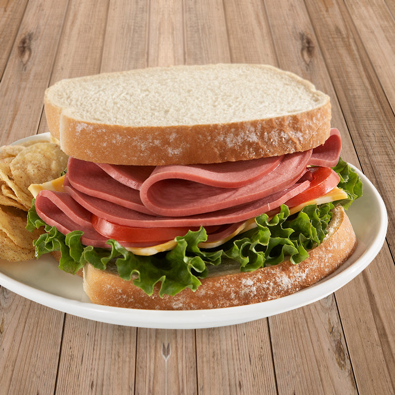A halal bologna sandwich on white bread with tomato, cheese, and lettuce. Chips are visible behind the sandwich to the left.