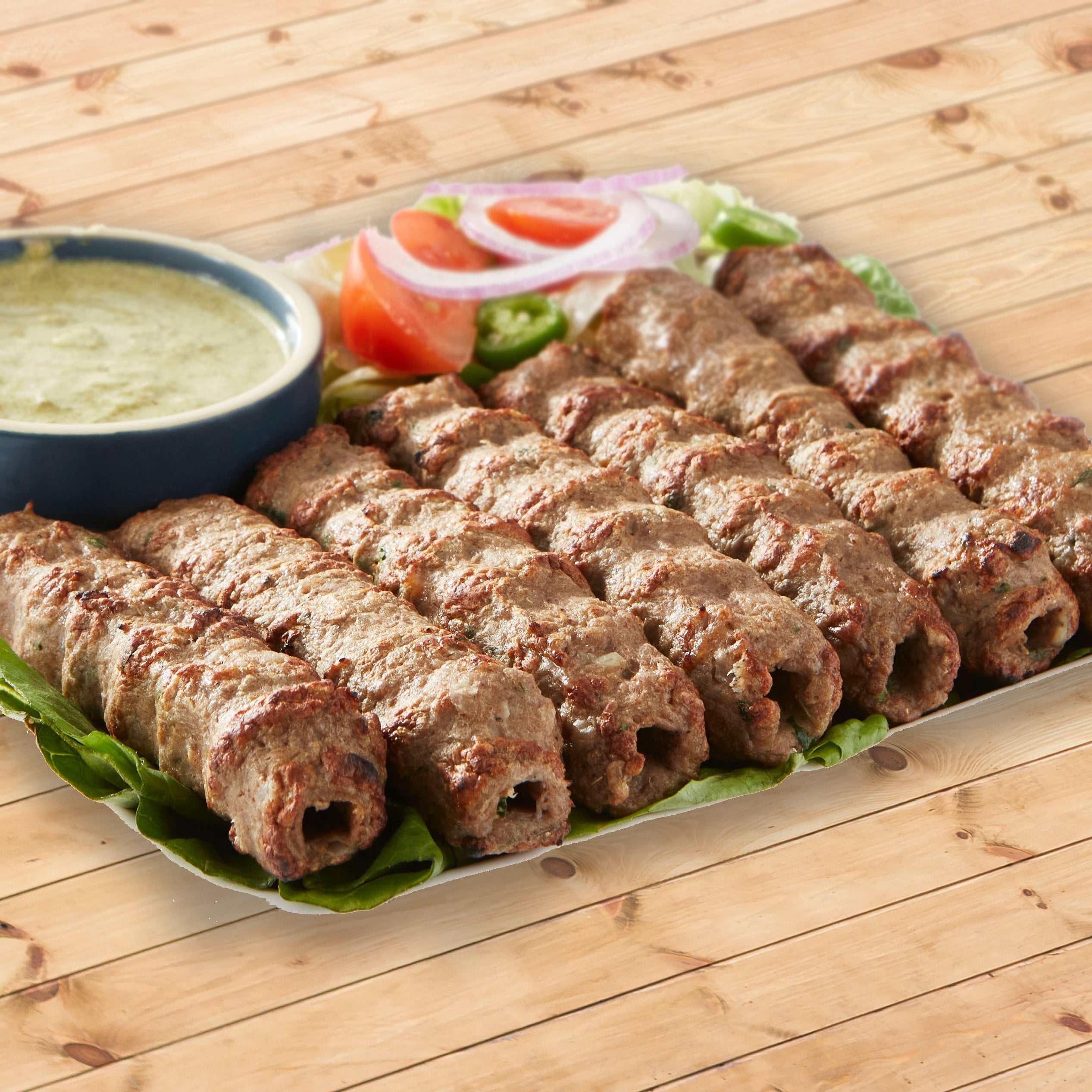 Beef seekh kabab resting on a bed of lettuce, it is garnished with onions and tomatoes, beside them is a blue bowl with a light green sauce