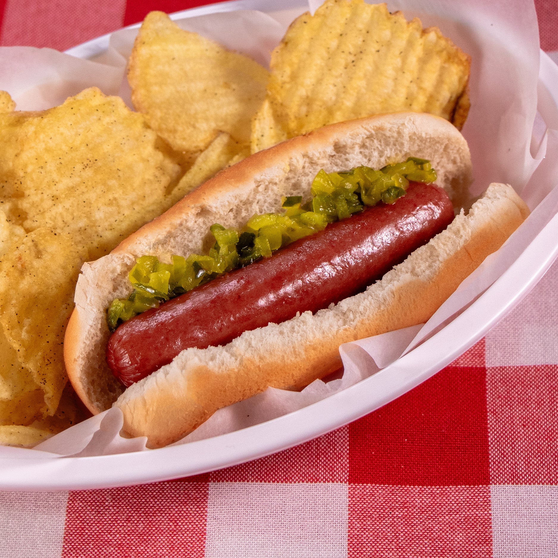 a Halal beef frank in a bun with pickle relish sits in a white basket with paper next to crinkle cut potato ships.