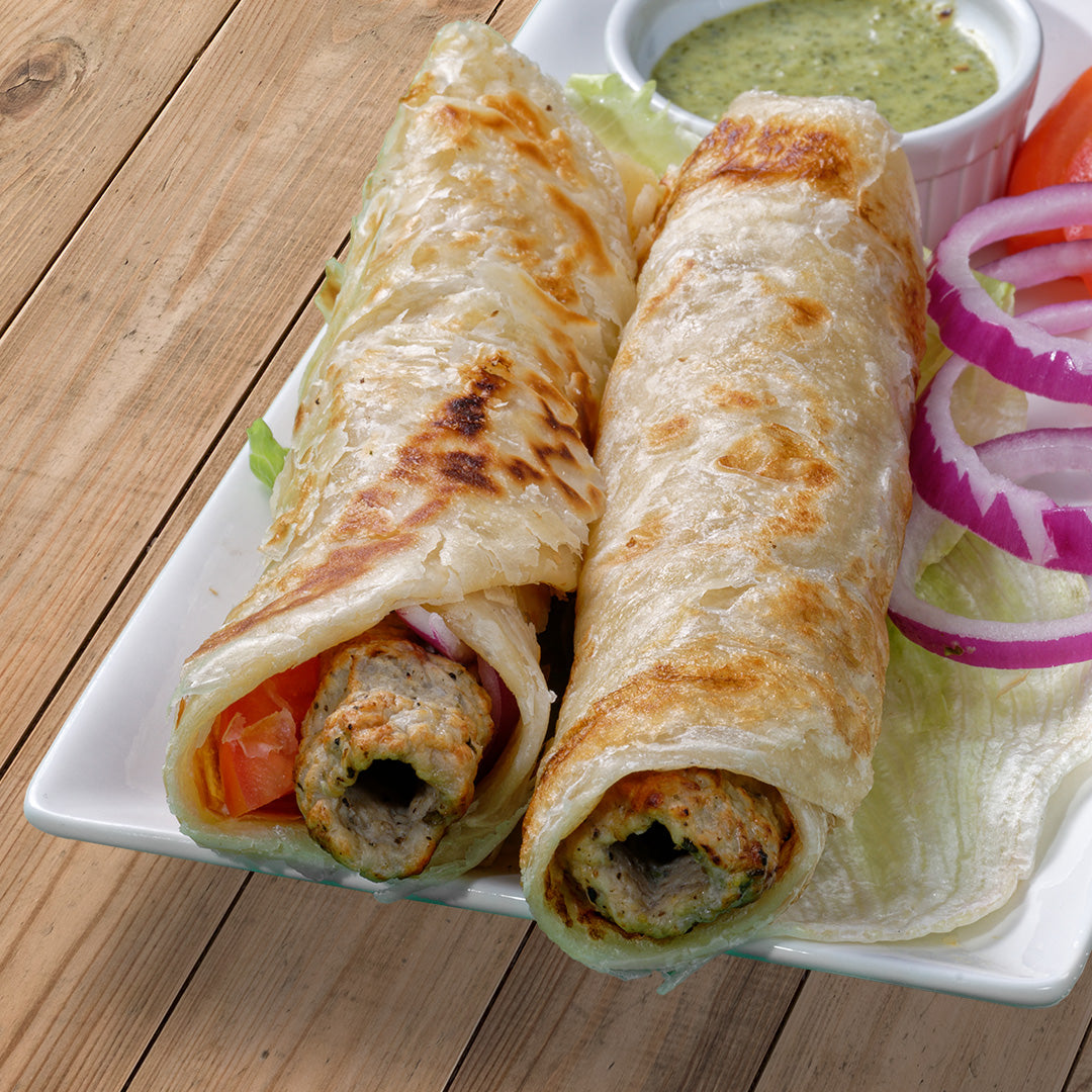 2 kati rolls made with chicken kebabs and wrapped in paratha are on a plate with a bowl of green chutney sitting behind them some rings of red onion are next to the rolls.