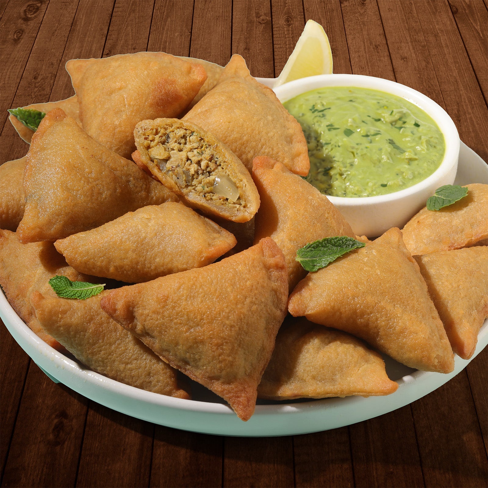 Kaiser Lahori Chicken Samosas in a pile on a white plate with some mint leaves. A bowl of chutney sits next to the pile. One samosa is cut in half and a minced meat mixture is visible inside.