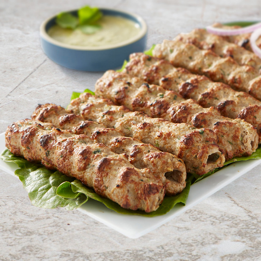 chicken seekh kababs resting leaves of lettuce. The food sits on a rectangular white plate. In the background sits a blow bowl with a beige sauce