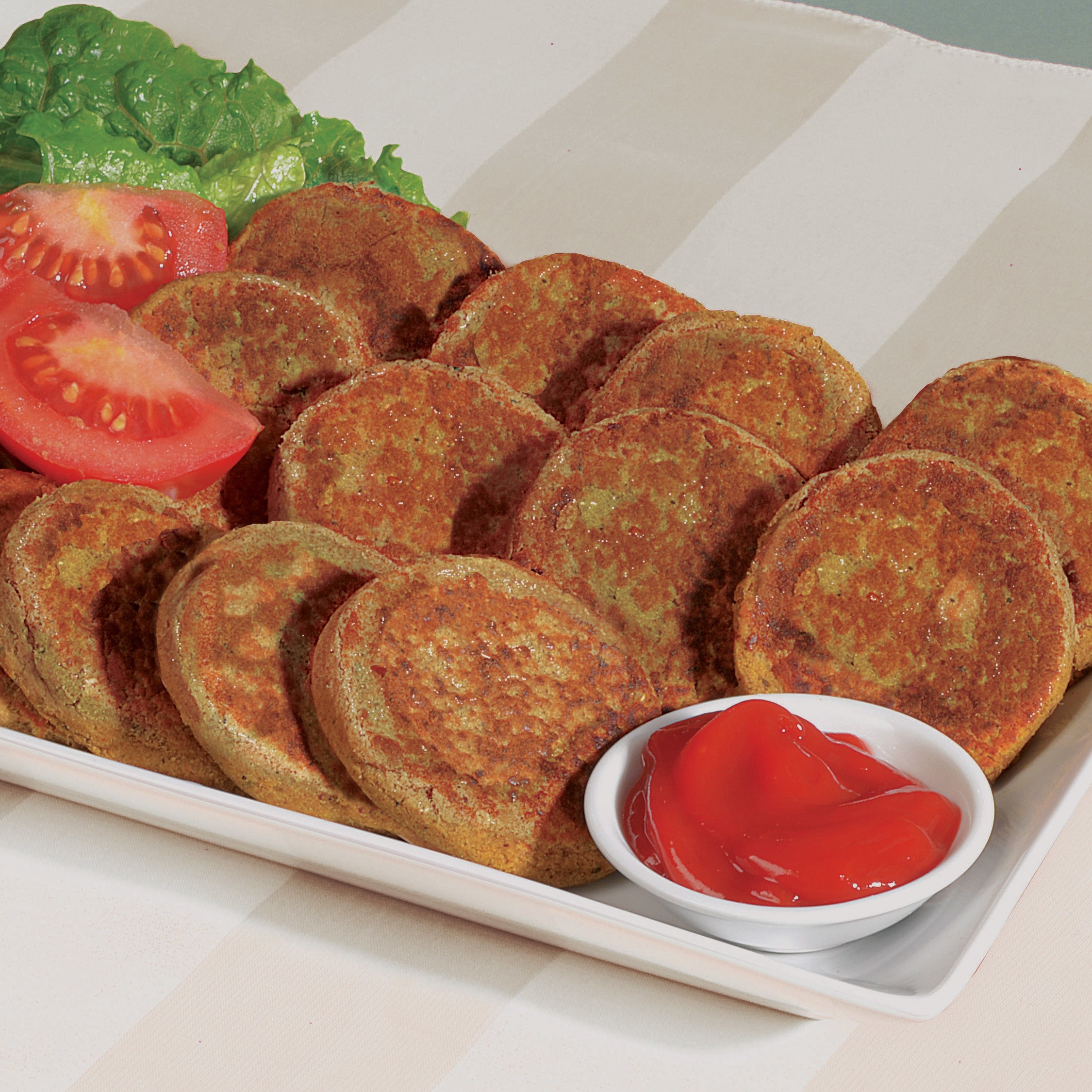 Chicken shami kababs sitting on a rectangular plate. On the back of the plate rest slices of tomato and a leaf of lettuce, on the front of the plate is small bowl with a red condiment.