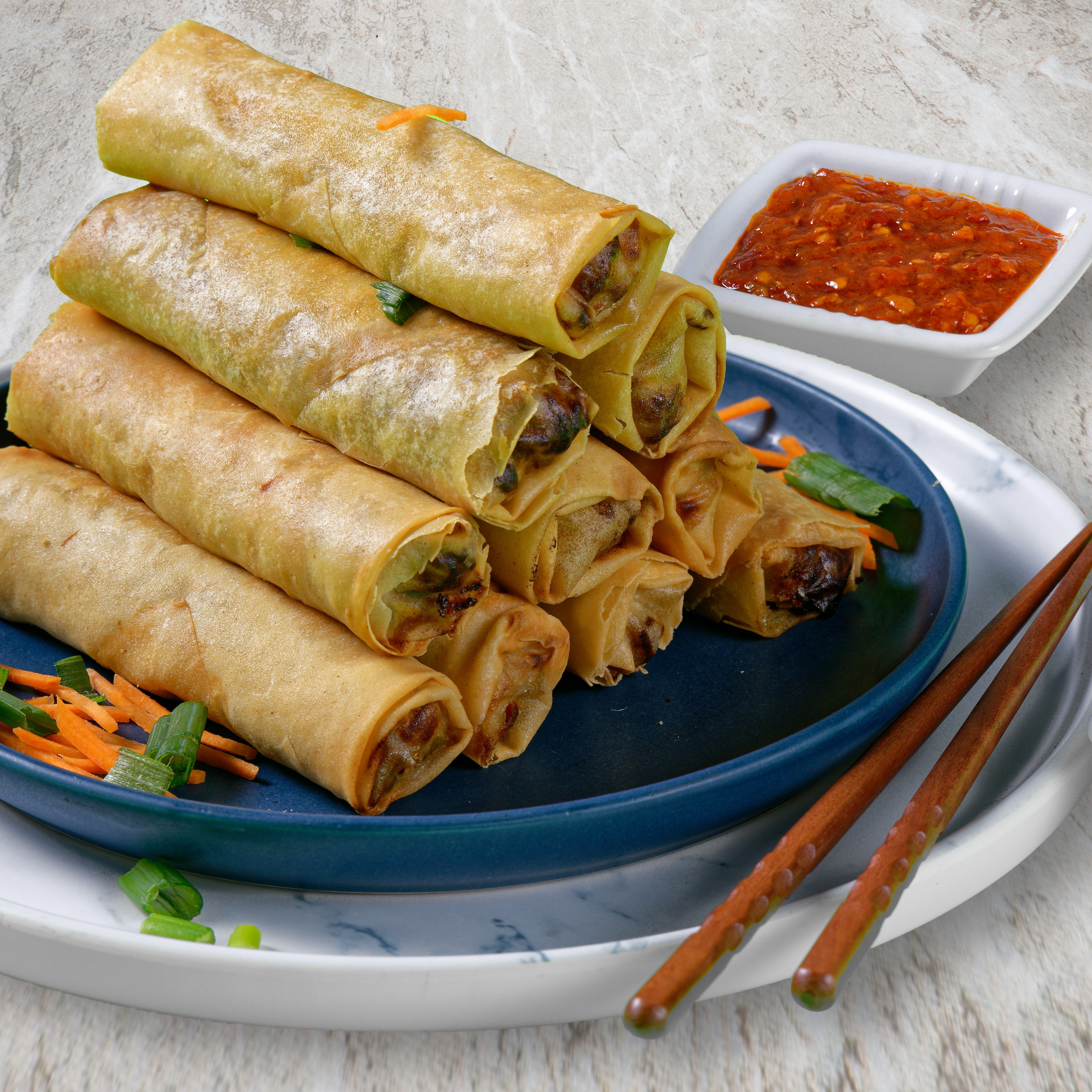 A pyramid of 10 crispy fried spring rolls sits on plate with a small pile of sliced carrots to the left. To the right and behind the pyramid is a small square bowl of chili paste.