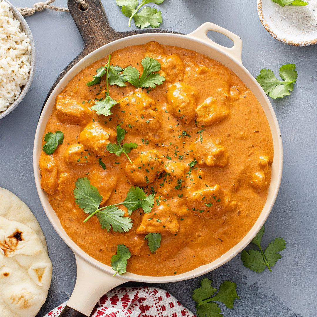 Chicken Tikka Masala with visible chunks of chicken in a ceramic pan. A bowl of rice and two pieces of naan are to the left of the pan. Cilantro is spread around the scene.