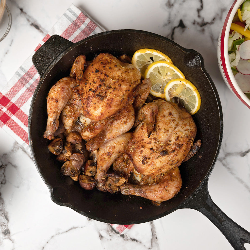2 cooked cornish hens in a cast iron pan with cooked vegetables and 3 slices of lemon. The pan rests on a patterned cloth on a marble counter top. Beside it is a bowl of salad