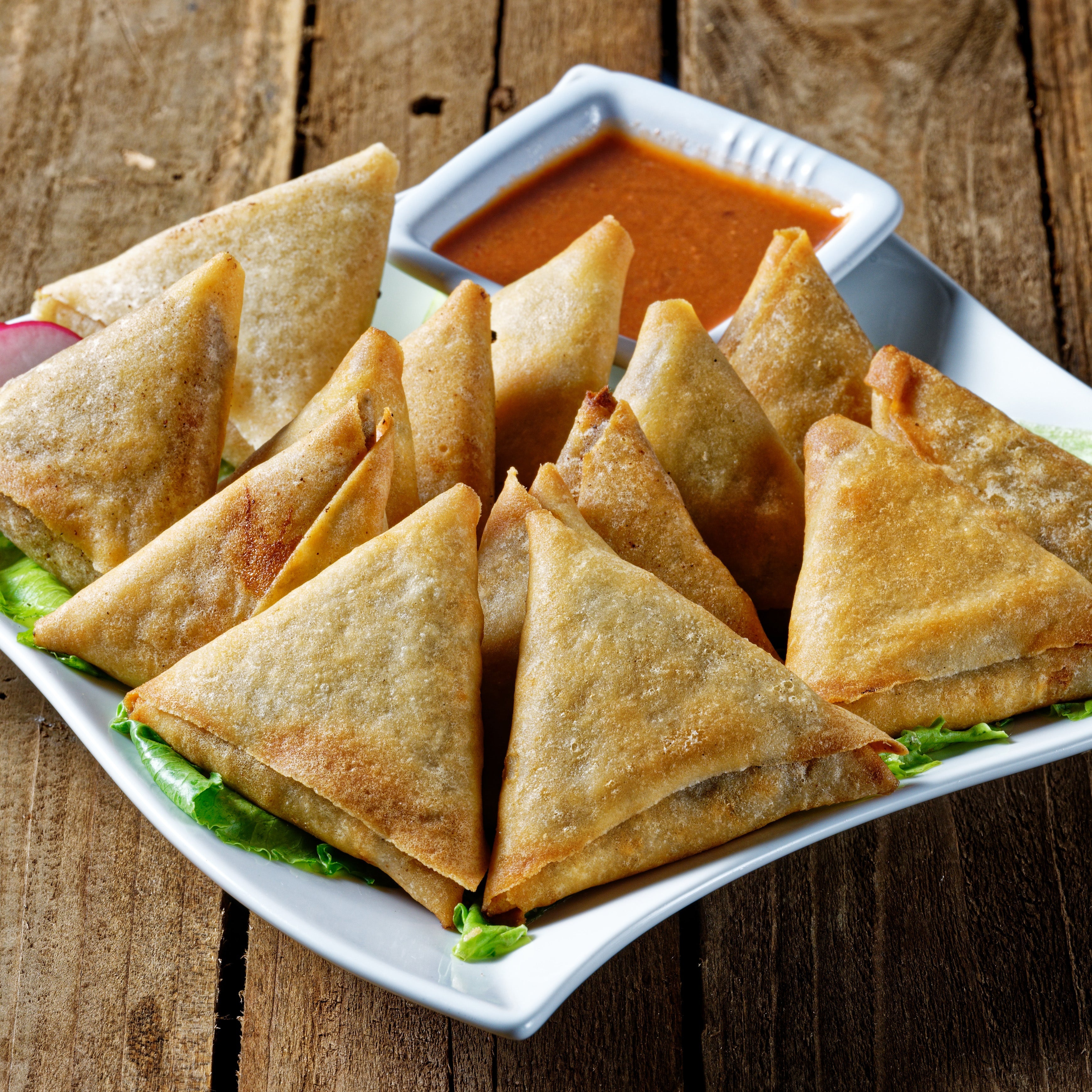 A white plate with crispy chicken samosas resting on a bed of lettuce. The samosas are garnished with a slice of radish and served with a bowl of reddish orange sauce