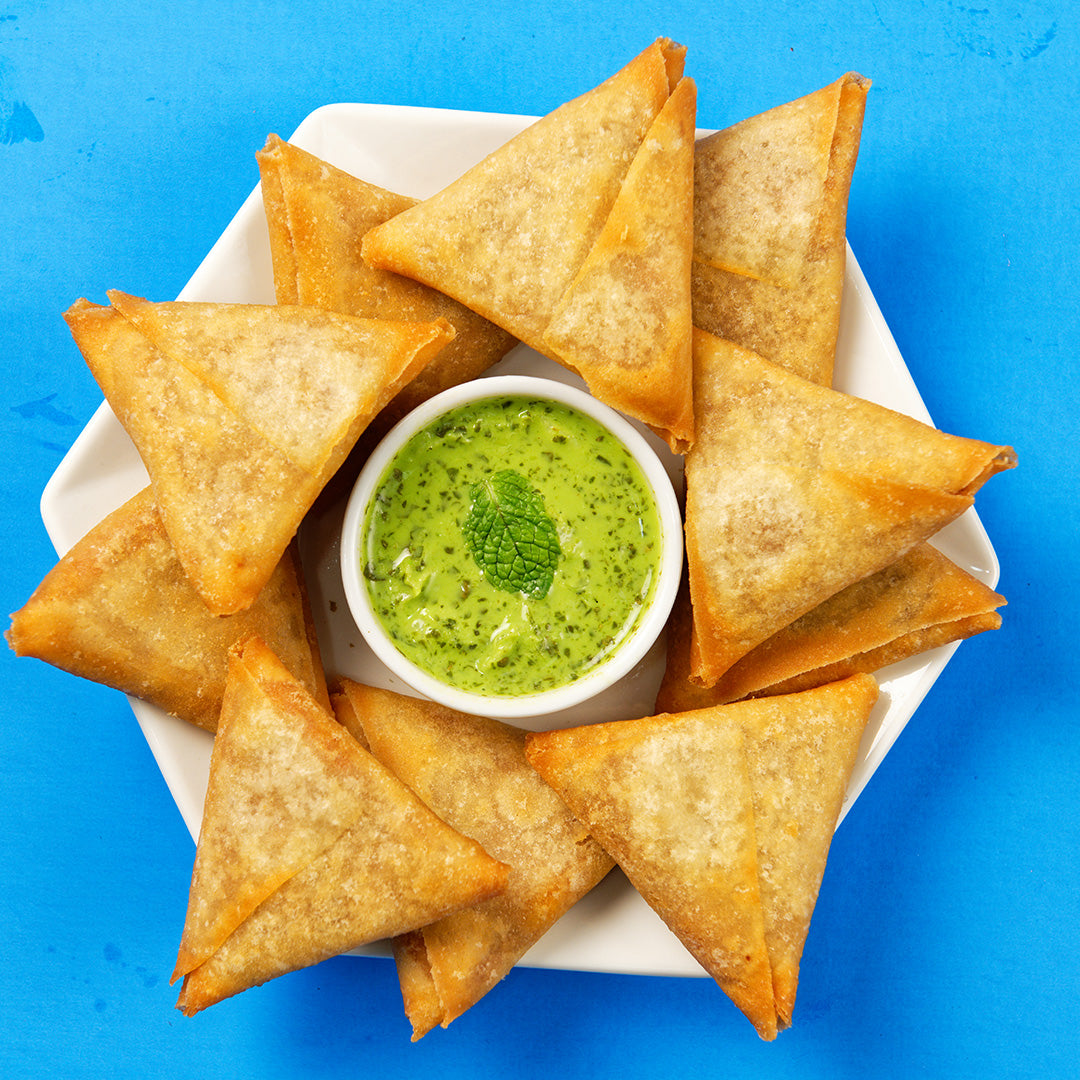 Chicken samosas layered on a white plate in a star pattern. In the center of the samosas is a green sauce with a single dark green leaf in the middle. The plate is resting on a blue background