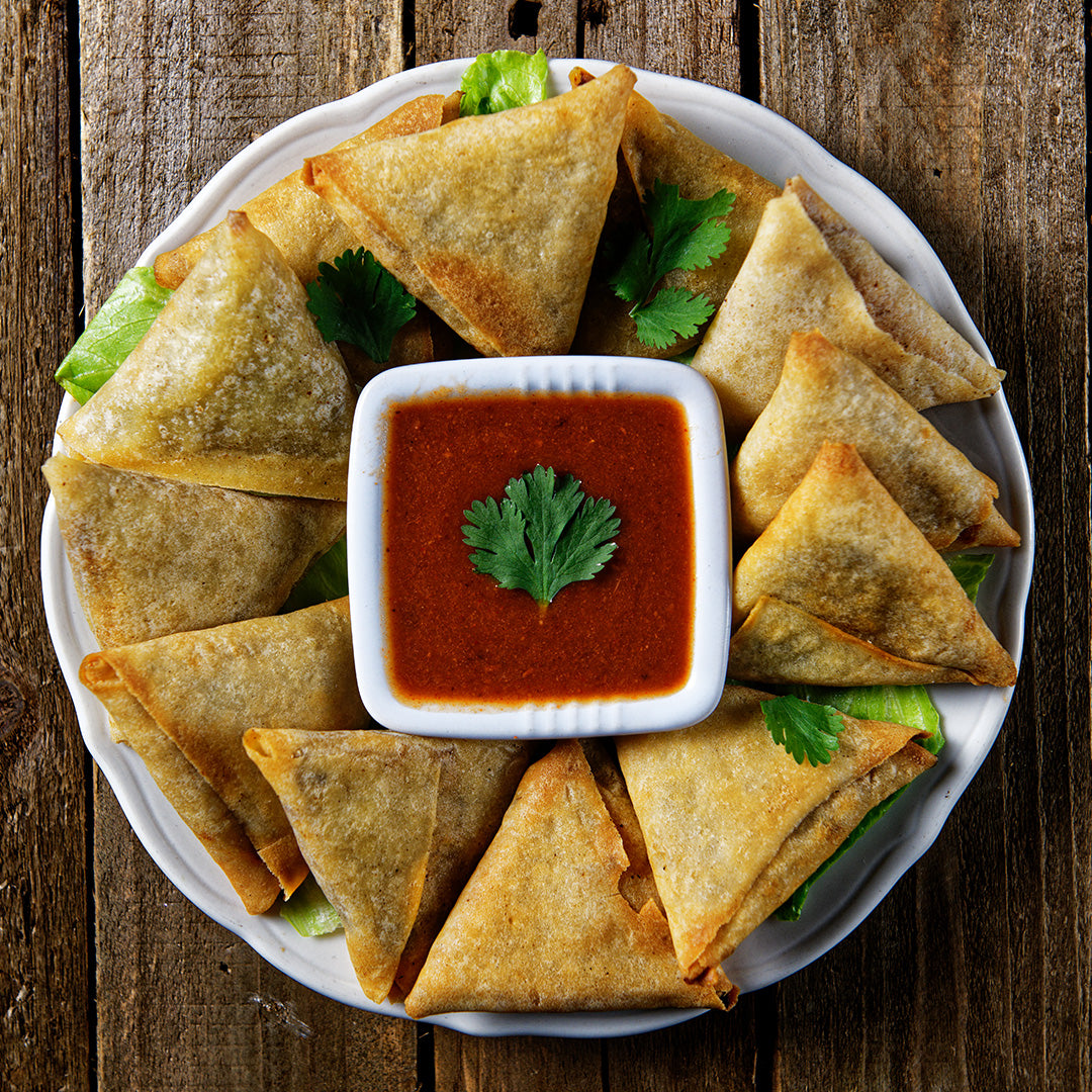 crispy lamb samosas arranged on a white plate, in the center is a square bowl with a red sauce. In the center of the sauce is a single leaf of cilantro, the rest of the plate is garnished with leaves of cilantro. The plate rests on a wooden background