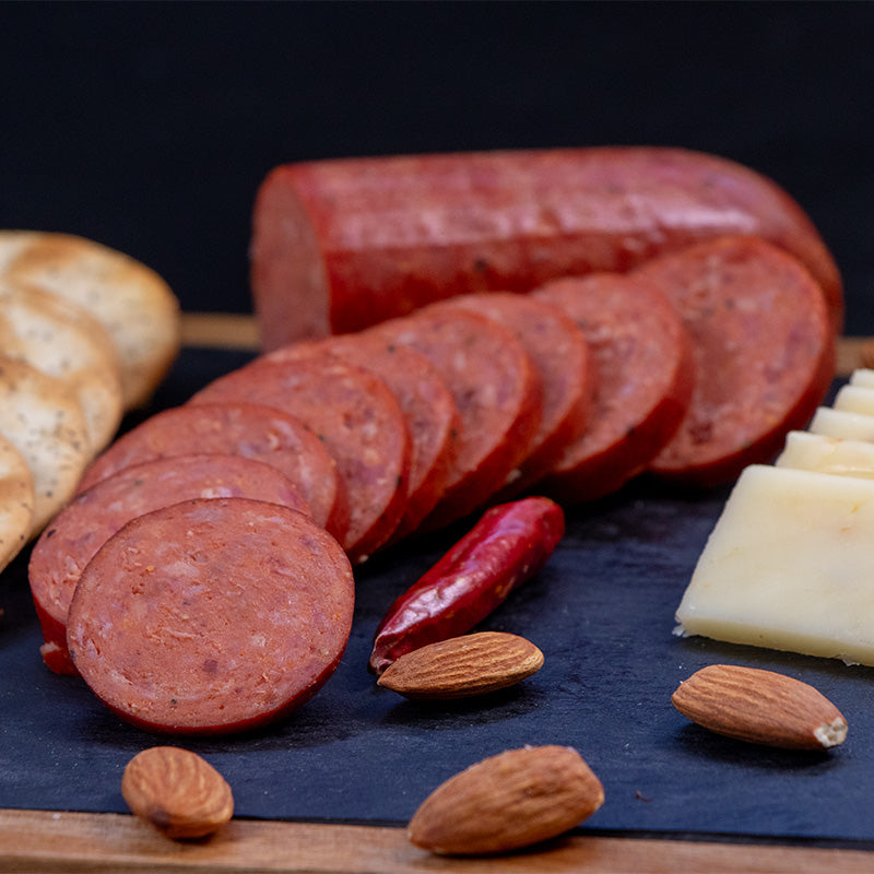 Slices of halal hot and spicy beef sausage is cut up and fanned out in a curve to the right. Large crackers are lined up to the left. A red dried pepper is to the right and some almonds are scattered around.