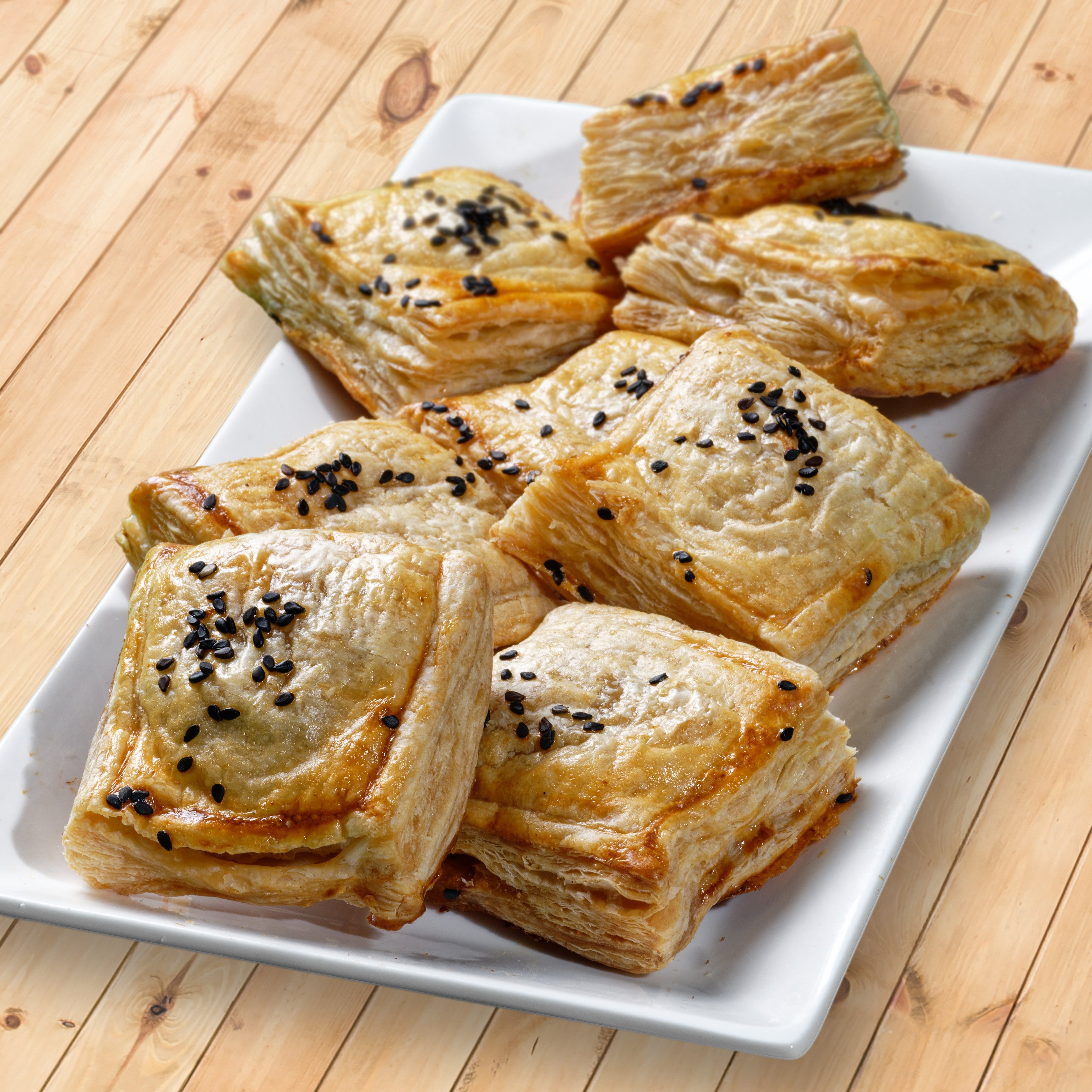 Eight square shaped puff pastry with visible flaky layers and topped with black sesame are arranged on a long rectangular plate.