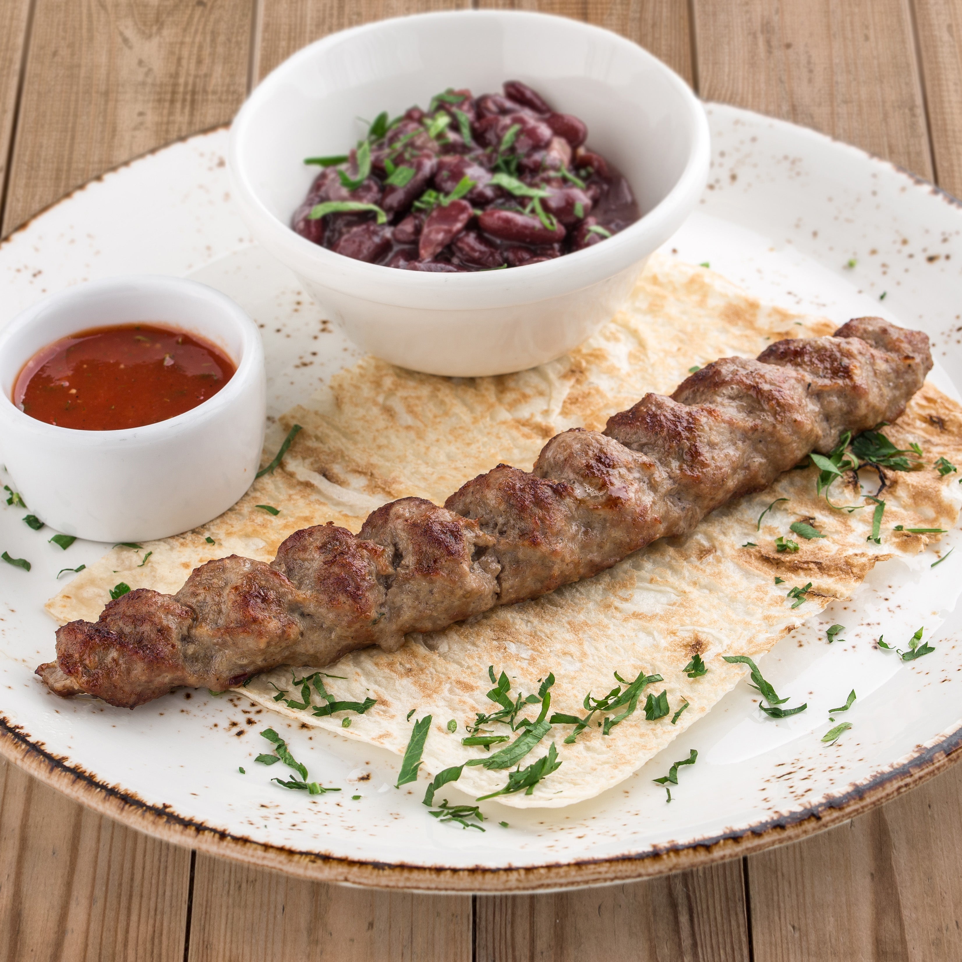 lamb kofta kabab resting on a thin piece of bread, beside it is small bowl with a red sauce and slightly larger white bowl with chopped olives. The product is garnished with chopped cilantro