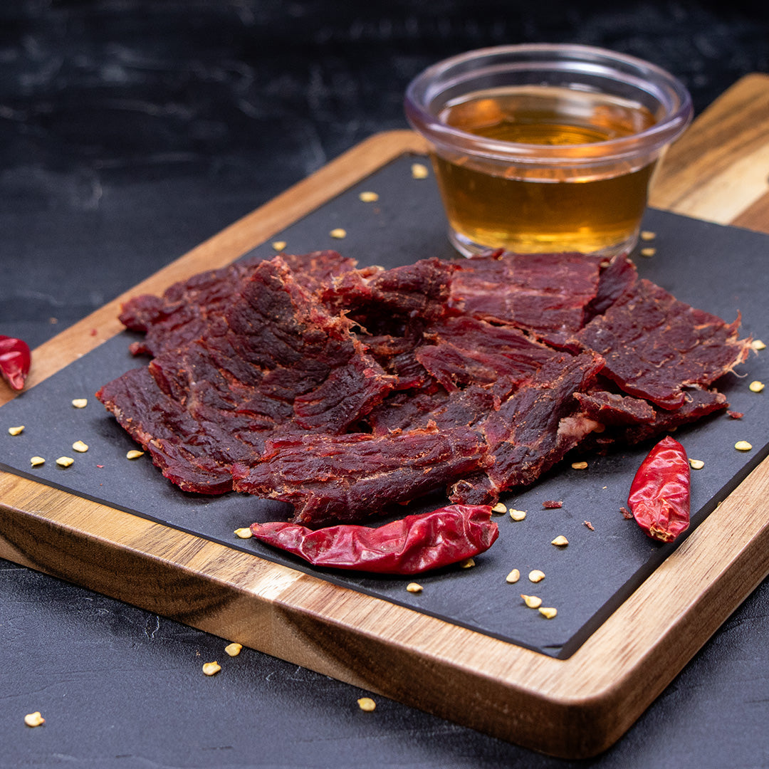 Midamar halal beef jerky strips sit on a cutting board surrounded by dried peppers and seeds, behind the jerky is a clear cup of honey