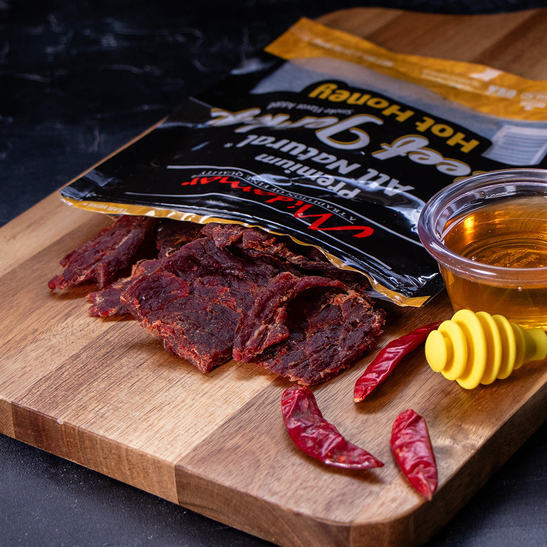 Midamar halal beef jerky pieces are spilling formm the bag on to a cutting board next to three dried peppers and a clear cup of honey