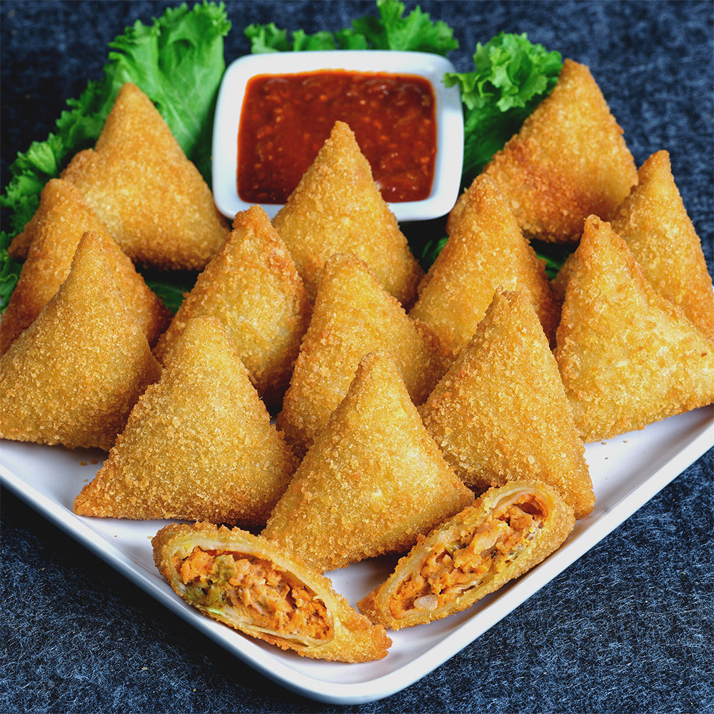 A white plate with 14 breaded chicken samosas arranged in line with one samosa cut open in the from exposing a minced chicken stuffing. a bed of lettuce in the back with a bowl of chili paste on it.