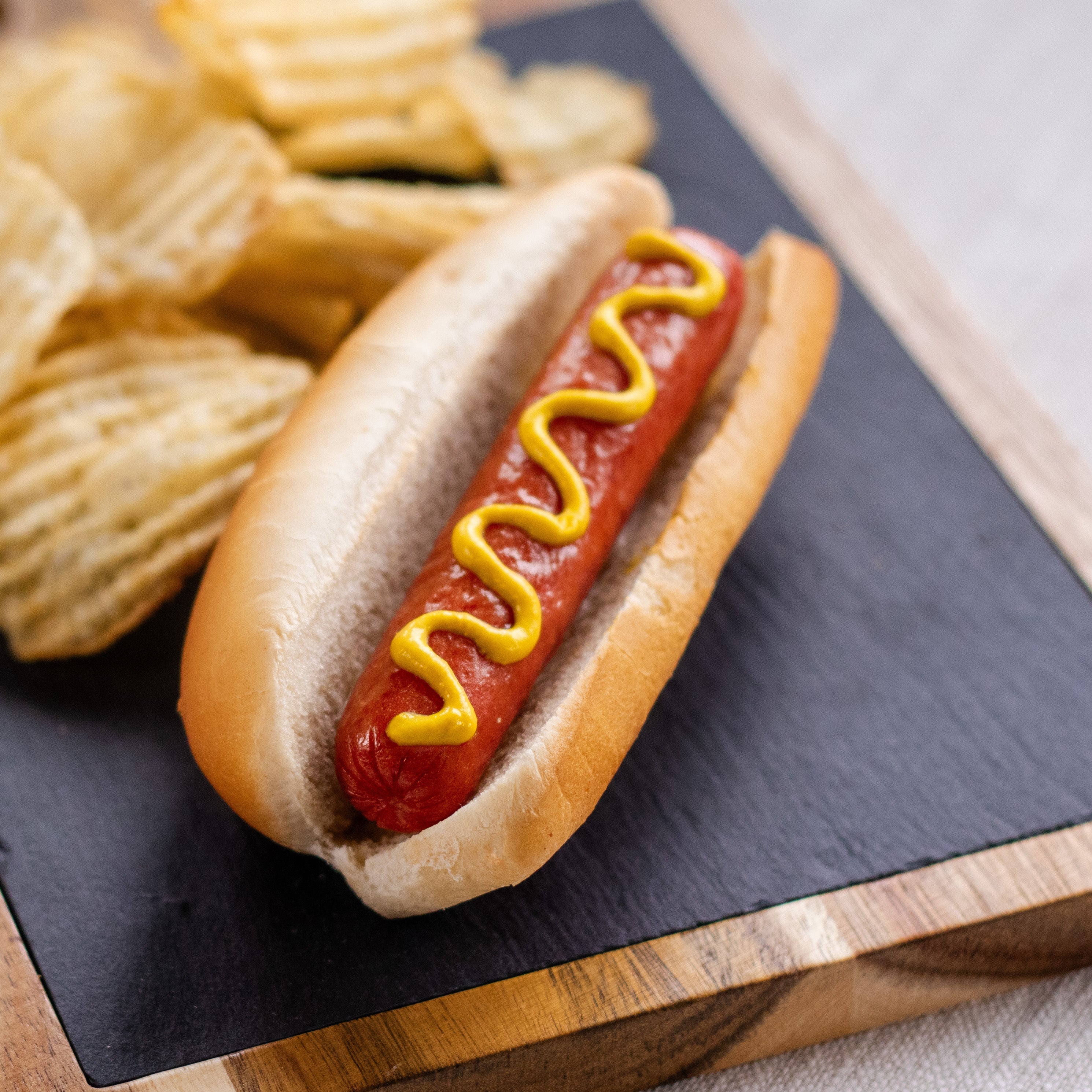 A halal beef frank in  a hot dog bun with mustard on it sits on a black cutting board with potato chips behind it.