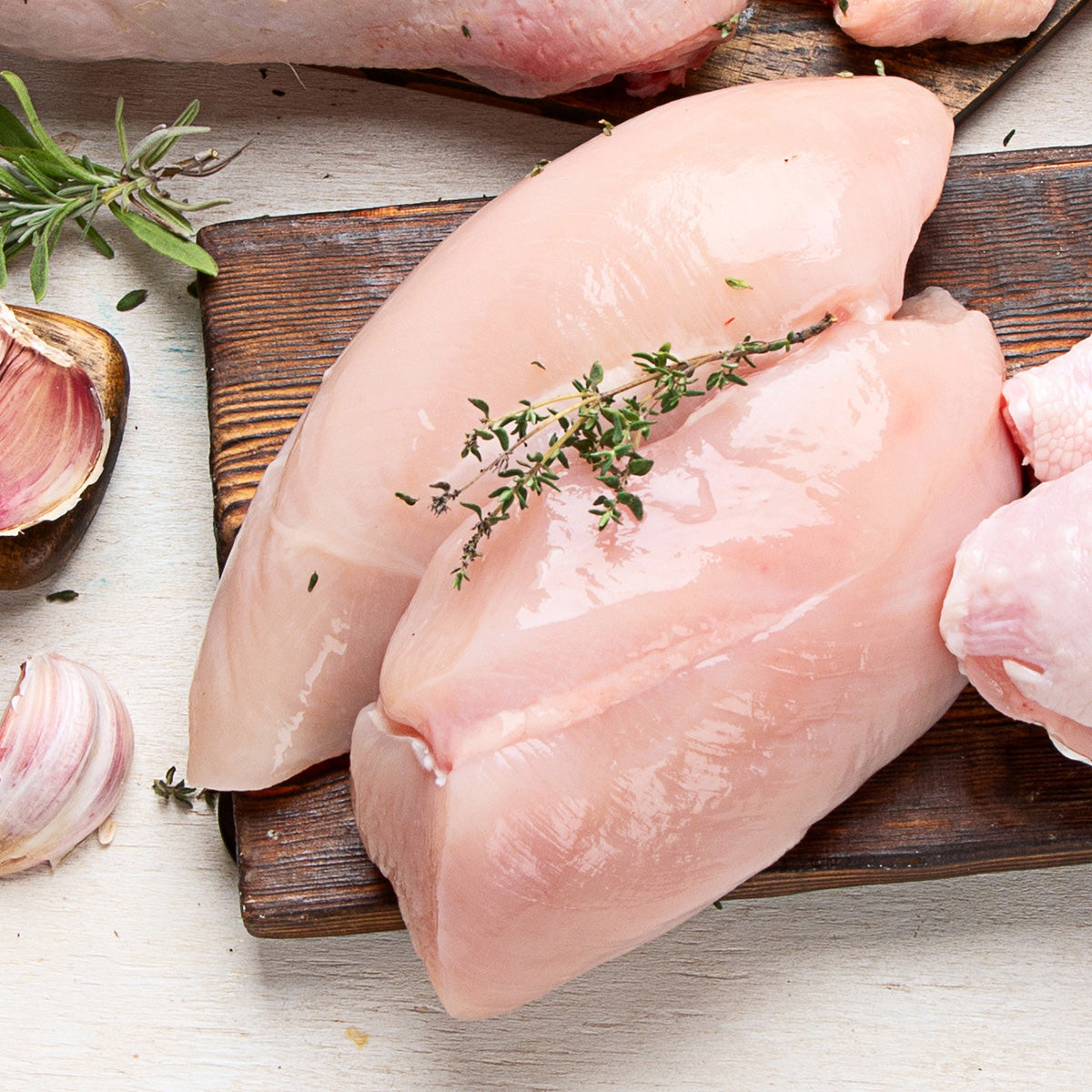 Raw chicken breasts resting on a dark wooden board, the chicken breast is garnished with a sprig of an herb